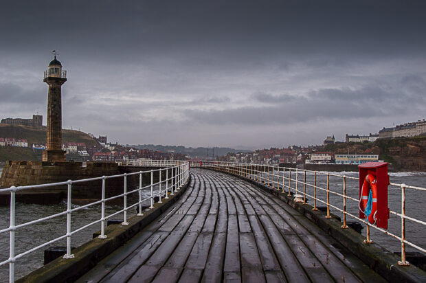 View from end of pier with lighthouse in the distance under grey sky.